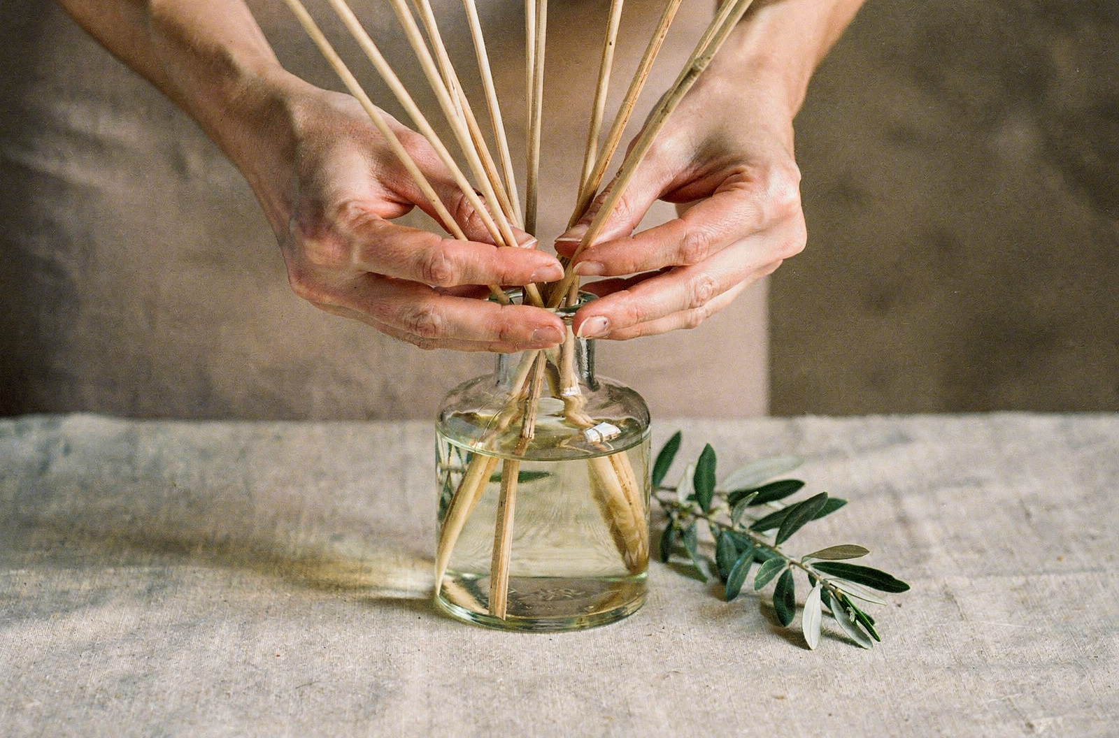 Hands arranging reed diffuser sticks in a glass bottle beside an olive sprig on linen